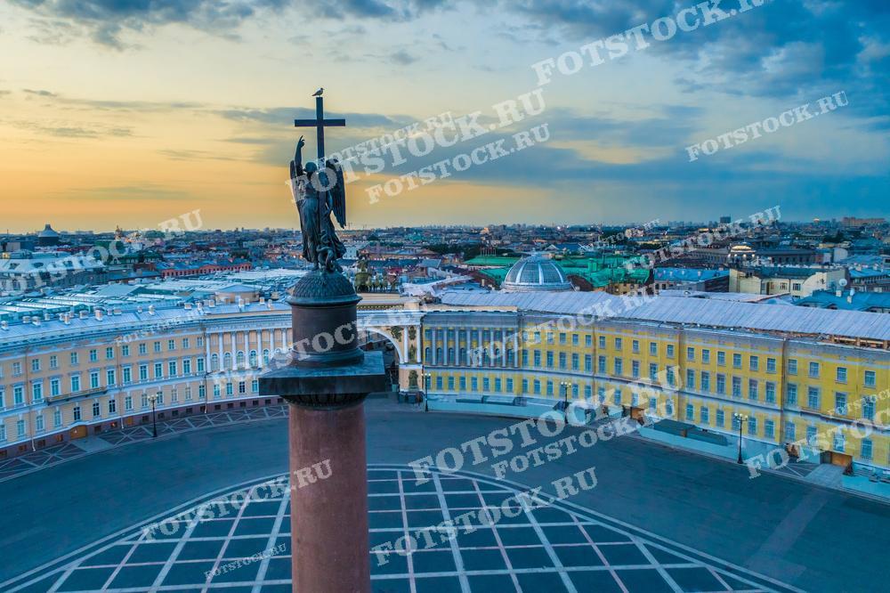 На фотографии изображён вечерний вид Дворцовой площади Санкт-Петербурга с высоты, видны крыши зданий и панорама города при закате. Это место идеально подходит для использования в туристических брошюрах, публикациях о культуре и истории города, а также может быть использовано для украшения статей о Санкт-Петербурге или как красивый фон для тематических презентаций.